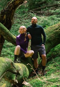 Uomo in piedi e donna seduta su un tronco d'albero caduto coperto di muschio in una foresta verde, entrambi indossano abbigliamento da escursionismo.