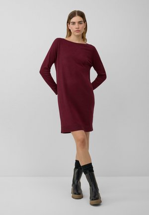 Robe en tricot bordeaux à manches longues, coupe décontractée, encolure ronde et poches latérales. Portée avec des bottines noires et des chaussettes texturées.