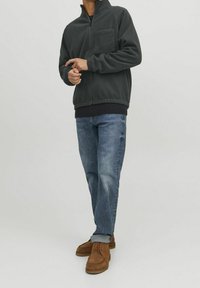 Tmavě šedá fleecová bunda na zip s stojáčkovým límcem, s náprsní kapsou a žebrovanými rukávy, v kombinaci s modrými džínami a hnědými botami.