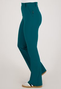 Vrouw in teal rechte broek en witte sneakers met zwarte strepen, staand tegen een effen lichte achtergrond.