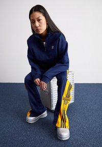 Dunkelblaues Zip-Hoodie mit kleinem Logo, kombiniert mit gelben und dunkelblauen Jogginghosen mit Streifen und Branding. Trägt klobige graue Schuhe.