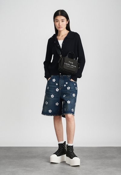 Sort strikket cardigan, hvid T-shirt, blå denim shorts med blomsterprint og sorte platformstøvler med snørebånd. Håndtaske med teksten: "DAGTASKE."