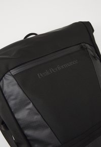 Black backpack with a textured surface, featuring a smooth panel, zippered pocket, and logo detail. Compact design with adjustable straps.