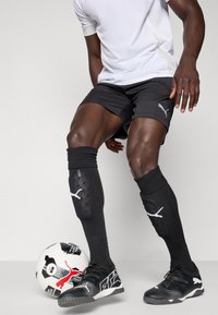Mustad musta Puma spordilühikesed püksid, valge dri-fit särk, mustad sääreguardid, mustad ja hallid spordijalatsid ning must-valge jalgpall.