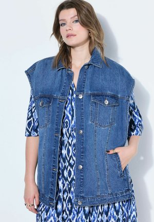 Kobieta w oversize'owej, bezrękawnikowej, niebieskiej kurtce dżinsowej na sobie niebieską sukienkę w wzory, z jedną ręką w kieszeni kurtki, stoi na tle białej ściany.