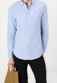 Man som bär en ljusblå skjorta med knappar och svarta byxor, håller en beige tygväska framför en vit bakgrund.