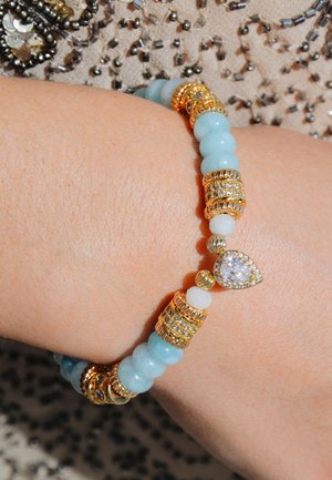 Un bracelet en perles avec des pierres bleu clair, des accents dorés et un gemme en forme de goutte d'eau claire. Le design comprend des perles rondes et en forme de baril alternées.