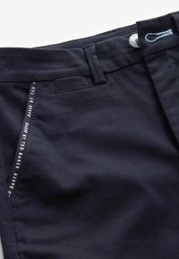 Pantaloni blu navy con una texture liscia, vestibilità regolare, tasche frontali e un'etichetta del marchio ben visibile lungo il bordo della tasca.