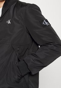 Czarna kurtka bomberka z zamkiem z przodu, ściągaczami na rękawach i naszywką z logo na rękawie. Wykonana z gładkiego, lekkiego materiału o matowym wykończeniu.