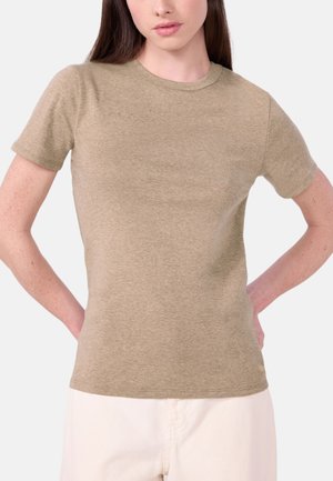 Frau trägt ein schlichtes beige Kurzarm-T-Shirt und helle Hosen, steht mit den Händen auf den Hüften.