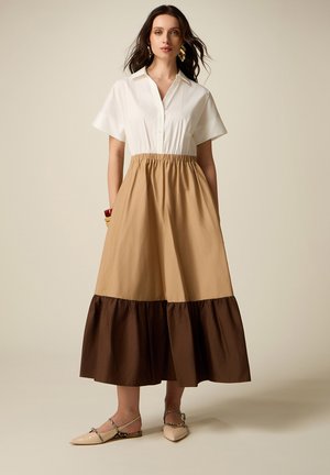 Vrouw draagt een wit blousejurkje met korte mouwen en kraag, een beige rok met donkerbruine geplooide zoom, gecombineerd met beige puntige schoenen.