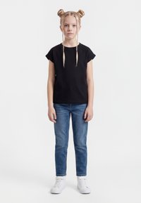 Ung jente med blondt hår i flettede boller, iført en svart T-skjorte, blå jeans og hvite sneakers, stående mot en ensfarget hvit bakgrunn.