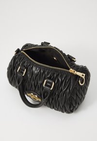 Schwarze gepolsterte Handtasche mit runder Form, goldenen Reißverschlüssen und Beschlägen. Innenansicht sichtbar, die ein Stofffutter zeigt. Zwei kurze Griffe sind enthalten.
