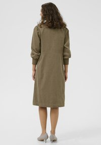 Olive green corduroy dress with long puff sleeves, fitted waist, and knee-length hem. Paired with gray heels, plain background.