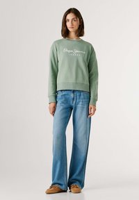 Φωτεινό πράσινο φούτερ με λευκό λογότυπο, συνδυασμένο με τζιν παντελόνια φαρδιάς γραμμής σε μπλε απόχρωση και καφέ παπούτσια σε στιλ μοκασίνι. Απλός σχεδιασμός και άνετη εφαρμογή.