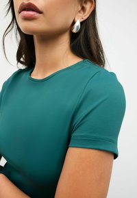 Frau in einem türkisfarbenen Kurzarmoberteil und silbernen tropfenförmigen sowie kleinen Steckerohrringen, mit dunklen Haaren vor einem einfarbigen Hintergrund.