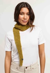 Une femme aux cheveux mi-longs et foncés porte une écharpe en laine jaune moutarde sur une chemise blanche à manches courtes et un pantalon beige, le tout sur un fond uni.