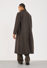 Long dark brown coat with a notched collar, textured fabric, single button cuffs, and side pockets, worn over loose-fitting gray pants.