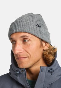 Grijze geribde beanie met een klein zwart patchlogo. Draag je met een grijze jas met een hoge kraag en zichtbare blauwe hardware details.