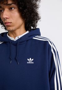 Marineblauwe hoodie met een witte drie-streep ontwerp en een klein wit Adidas-logo op de borst. Gemaakt van een zachte, gestructureerde stof.