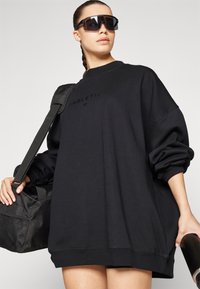 Sudadera negra de gran tamaño con mangas largas, cuello redondo y un sutil detalle del logo en el centro. La modelo sostiene un bolso de mano negro.