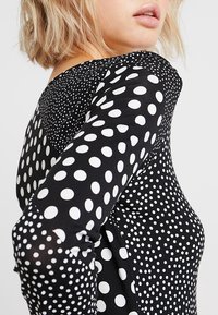 Haut à manches longues avec une base noire et des pois blancs de tailles variées. Texture de tissu douce avec un col rond et une silhouette ajustée.
