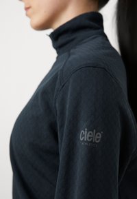 Temno mornarsko modra športna majica z visokim ovratnikom, teksturiranim vzorcem in diskretno oznako na roki, zasnovana za udobje in zmogljivost.