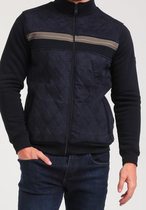 Mann trägt eine marineblaue, gesteppte Jacke mit Reißverschluss und einem beigen sowie schwarzen horizontalen Streifen über der Brust, dunkelblaue Jeans, eine Hand in der Tasche.