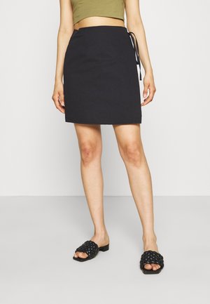 Personne portant une jupe portefeuille noire nouée à la taille, un crop top vert olive et des sandales tissées noires, debout sur un sol gris clair.