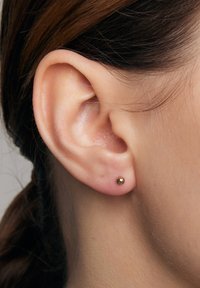 Mała złota kolczyk w przekłutym uchu. Gładka, okrągła metalowa powierzchnia o wypolerowanym wykończeniu, umieszczona centralnie na płatku ucha.