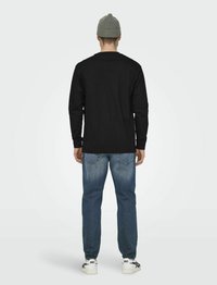 Schwarzes Langarmshirt, blaue ausgebleichte Jeans und weiße Sneakers mit schwarzen Akzenten. Graue Strickmütze auf dem Kopf. Neutraler Hintergrund.