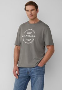 Grijze T-shirt met korte mouwen, featuring een ronde witte grafische tekstontwerp. Gepaard met lichtblauwe jeans, die een casual, relaxte pasvorm tonen.