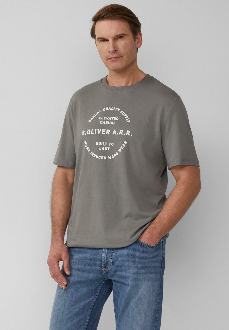 Grijze T-shirt met korte mouwen, featuring een ronde witte grafische tekstontwerp. Gepaard met lichtblauwe jeans, die een casual, relaxte pasvorm tonen.