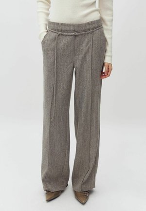 Femme portant un pantalon gris en tweed à taille haute et jambes larges avec une ceinture à cordon, associé à un pull côtelé blanc et des chaussures pointues marron.