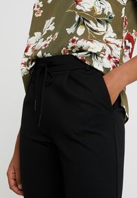 Pantalons noirs fuselés avec une taille à cordon, associés à un haut à imprimé floral vert et blanc, à manches courtes.