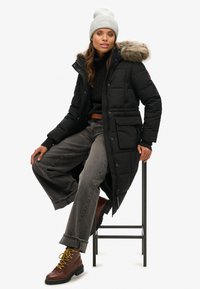 Abrigo acolchado negro con capucha de piel sintética, combinado con un suéter de punto gris, jeans de pierna ancha grises y botas marrones con cordones amarillos.