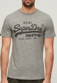 Camiseta de algodón gris con un logo impreso en negro y texto estilizado. Mangas cortas y cuello redondo. Textura mínima, diseño casual.