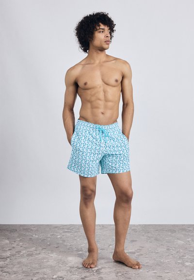 Muž s kučeravými vlasmi, ktorý má na sebe modré plavky s vzorom, stojí bosý na šedej kamennej podlahe, hľadí do strany s rukami v vreckách.