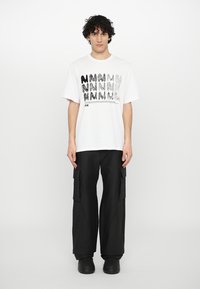 T-shirt bianca con stampa grafica nera di lettere stilizzate, abbinata a pantaloni cargo neri larghi e zoccoli neri.