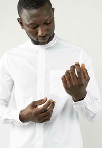 Weißes Hemd mit Stehkragen, Brusttasche und langen Ärmeln. Modell passt mit beiden Händen die Manschetten an. Glatte Stofftextur.