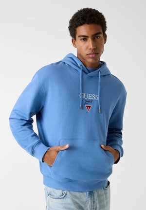 Jonge man die een lichtblauwe GUESS hoodie draagt met handen in de buidelzak, gecombineerd met lichtgewassen spijkerbroek, staand tegen een effen achtergrond.