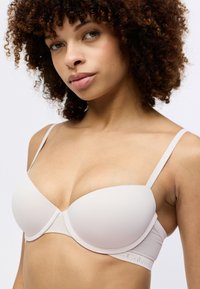 Beżowy stanik wyściełany, z regulowanymi ramiączkami, gładką fakturą, przezroczystymi panelami bocznymi oraz logo Calvin Klein na pasku. Subtelne detale w designie.