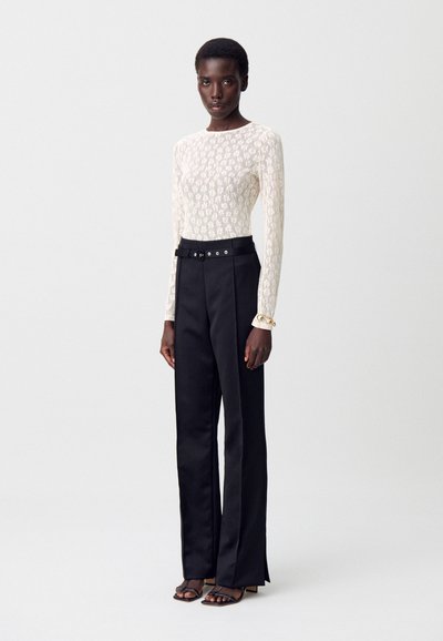 Mujer de pie con una blusa blanca de encaje floral de manga larga, pantalones negros de pierna ancha y cintura alta con un cinturón, y sandalias negras.