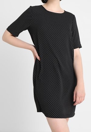 Donna che indossa un vestito nero corto con pois bianchi, una mano sul fianco e l'altra lungo il corpo, su sfondo semplice.