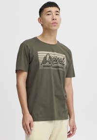Tricou verde măsliniu dintr-un material moale, cu un print grafic în bej și negru, având textul „Original” și linii orizontale. Mâneci scurte.