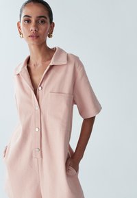 Svetlo roza kombinezon s kratkimi rokavi, gumbi spredaj in žepom na prsih. Iz teksturiranega materiala, s klasičnim ovratnikom in stranskimi žepi.