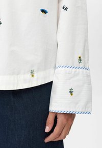 Witte lange mouwen shirt met kleine geborduurde bloemenpatronen en blauw gestreepte manchetten, gedragen over een donkere spijkerbroek.