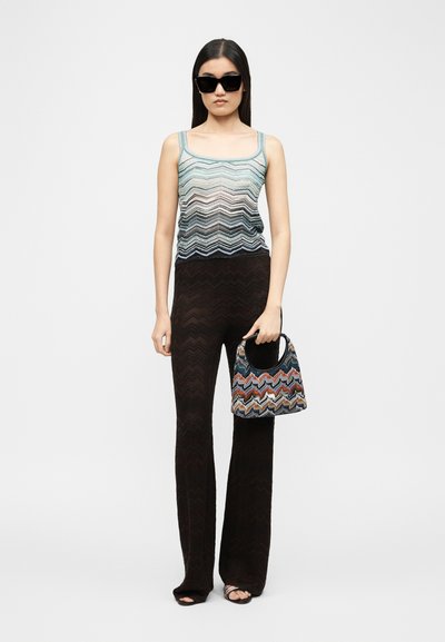 Γυναίκα με αμάνικη μπλούζα με σχέδια, μαύρο παντελόνι φαρδιάς γραμμής με υφή, μαύρα γυαλιά ηλίου, κρατώντας πολύχρωμη τσάντα με ζιγκ-ζαγκ μοτίβο.