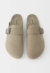 Sabots en daim beige avec une large sangle ornée d'une boucle en métal argenté, bout rond et dessus texturé. Taille 45 indiquée à l'intérieur de la chaussure.