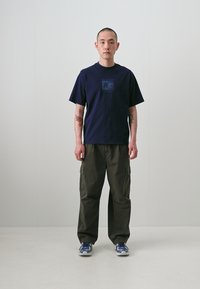 T-shirt en coton bleu marine avec un imprimé graphique sur la poitrine, associé à un pantalon cargo olive et des chaussures de sport bleues avec des accents blancs.